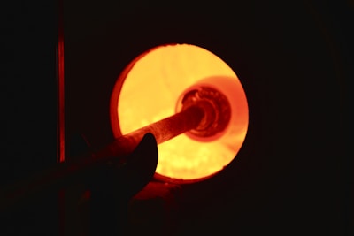 A glowing hot furnace with an intense orange light emanating from within. A metal tool is being inserted into the furnace, possibly for a glassblowing or metalworking process.
