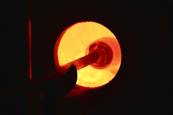 A glowing hot furnace with an intense orange light emanating from within. A metal tool is being inserted into the furnace, possibly for a glassblowing or metalworking process.