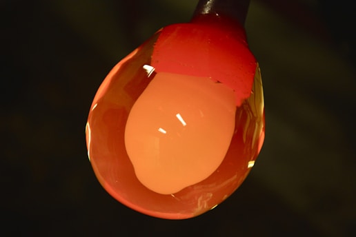 A close-up view of molten glass being shaped, with a glowing orange color. The glass has a bulbous form and appears shiny with reflective surfaces.