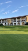 A golf course is seen with a well-maintained green area and a flagstick. In the background, a two-story building with a stone and stucco facade features several archways and balconies. A clear blue sky enhances the serene setting.