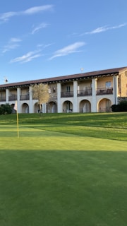 A golf course is seen with a well-maintained green area and a flagstick. In the background, a two-story building with a stone and stucco facade features several archways and balconies. A clear blue sky enhances the serene setting.