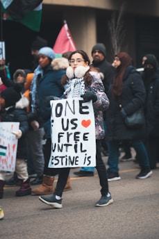 A group of people are participating in a protest march. A young person in the foreground wears earmuffs and glasses, holding a sign that reads 'None of us are free until Palestine is.' The crowd is dressed warmly, suggesting cold weather. Flags can be seen being held by other participants.