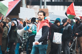 A group of people gathered in a protest or rally, with individuals holding signs and Palestinian flags. The central figure is holding a megaphone and wearing a jacket with symbolic colors. The crowd in the background is diverse and engaged in the demonstration.