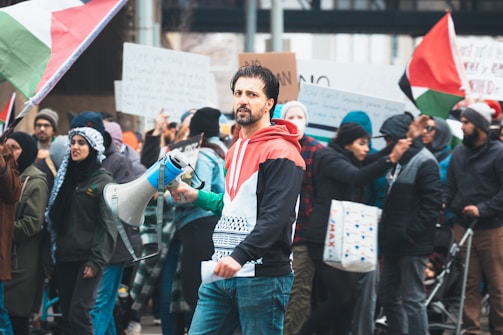 A group of people gathered in a protest or rally, with individuals holding signs and Palestinian flags. The central figure is holding a megaphone and wearing a jacket with symbolic colors. The crowd in the background is diverse and engaged in the demonstration.
