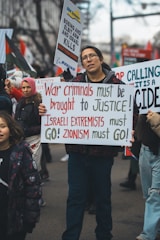 People are participating in a street protest. A man at the forefront holds a sign that reads 'War criminals must be brought to justice! Israeli extremists must go! Zionism must go!' Other protestors behind him also hold different signs, some with texts against Boeing and Elbit Systems, and messages calling for justice.