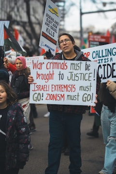 People are participating in a street protest. A man at the forefront holds a sign that reads 'War criminals must be brought to justice! Israeli extremists must go! Zionism must go!' Other protestors behind him also hold different signs, some with texts against Boeing and Elbit Systems, and messages calling for justice.