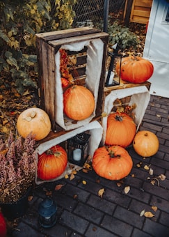 A rustic autumn display featuring several orange pumpkins arranged on and around textured wooden crates. The setting includes lanterns with candles and autumn leaves scattered on a cobblestone surface. A pot with fall-blooming heather adds to the seasonal atmosphere.