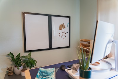 A workspace featuring a dual-panel whiteboard mounted on a light-colored wall. The right panel contains magnets and a small tool or pen. The desk has a computer monitor, a keyboard, a black mouse, a notebook with a blue cover, and an arrangement of potted plants including a tall thin plant and a leafy one. To the right, there is a wooden letter tray with papers.