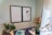 A workspace featuring a dual-panel whiteboard mounted on a light-colored wall. The right panel contains magnets and a small tool or pen. The desk has a computer monitor, a keyboard, a black mouse, a notebook with a blue cover, and an arrangement of potted plants including a tall thin plant and a leafy one. To the right, there is a wooden letter tray with papers.