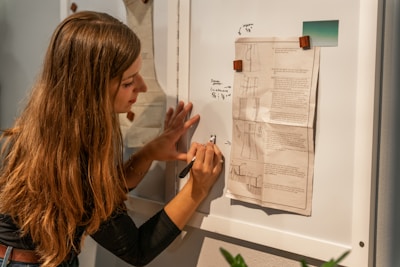 A person with long hair is writing notes on a whiteboard. Papers with diagrams and textual annotations are pinned to the board using wooden clips. The person appears focused and is using a pen to make annotations.