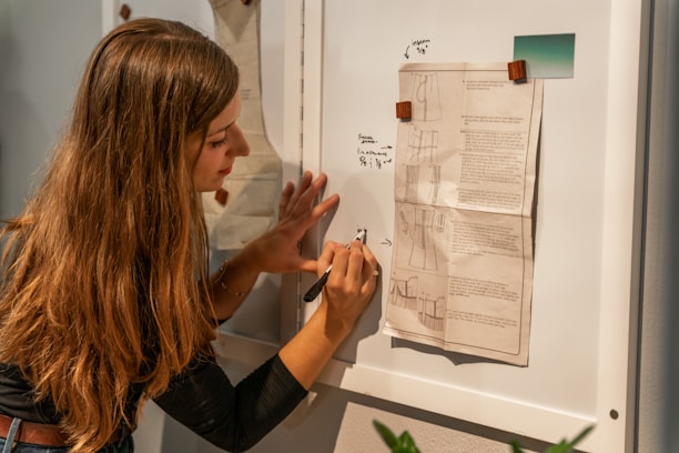 A person with long hair is writing notes on a whiteboard. Papers with diagrams and textual annotations are pinned to the board using wooden clips. The person appears focused and is using a pen to make annotations.