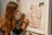 A person with long hair is writing notes on a whiteboard. Papers with diagrams and textual annotations are pinned to the board using wooden clips. The person appears focused and is using a pen to make annotations.