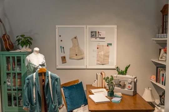A cozy sewing room featuring a dress mannequin draped with fabric, a light teal jacket on a coat hanger, and a sewing machine on a wooden desk. The desk is adorned with a potted plant, open notebook, and various sewing supplies. A green cabinet on the left holds a ukulele and a plant on top. Two framed sewing patterns are mounted on the wall behind the desk, with books neatly arranged beside the sewing machine. Shelves in the background contain books and decorative items.