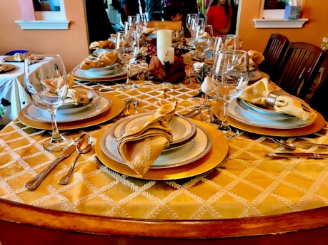 Elegant dining table set with fine tableware, featuring beautifully arranged plates, glasses, and folded napkins on a golden patterned tablecloth. The table is adorned with silverware, and a decorative centerpiece including a candle and autumn-themed elements. Surrounding chairs suggest a welcoming setting for a meal.