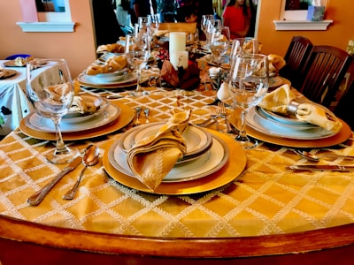 Elegant dining table set with fine tableware, featuring beautifully arranged plates, glasses, and folded napkins on a golden patterned tablecloth. The table is adorned with silverware, and a decorative centerpiece including a candle and autumn-themed elements. Surrounding chairs suggest a welcoming setting for a meal.