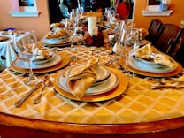 Elegant dining table set with fine tableware, featuring beautifully arranged plates, glasses, and folded napkins on a golden patterned tablecloth. The table is adorned with silverware, and a decorative centerpiece including a candle and autumn-themed elements. Surrounding chairs suggest a welcoming setting for a meal.