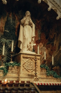 A marble statue depicting a religious figure dressed in robes, positioned on a decorated altar adorned with ornate carvings. The altar is flanked by lit candles and surrounded by greenery. The backdrop features a dark mosaic with intricate patterns.