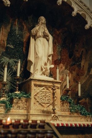 A marble statue depicting a religious figure dressed in robes, positioned on a decorated altar adorned with ornate carvings. The altar is flanked by lit candles and surrounded by greenery. The backdrop features a dark mosaic with intricate patterns.