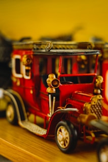 A detailed, vintage-style model car in bright red with gold accents. It features ornate details, including a grill and headlights. The vehicle is set against a blurred background, highlighting its intricate craftsmanship.