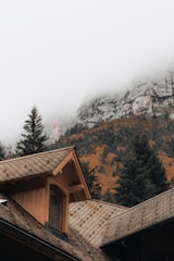 A charming wooden cottage nestled among tall pine trees with misty Himalayan peaks in the background.