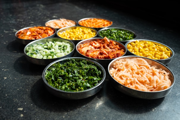 A variety of colorful vegetables and pickled or seasoned foods are arranged in small metal bowls on a dark surface. Each bowl contains a different type of food, such as leafy greens, corn, diced cucumbers, kimchi, and pickled radishes, showcasing a mix of textures and colors.