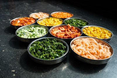 A variety of colorful vegetables and pickled or seasoned foods are arranged in small metal bowls on a dark surface. Each bowl contains a different type of food, such as leafy greens, corn, diced cucumbers, kimchi, and pickled radishes, showcasing a mix of textures and colors.