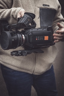 A person is holding a professional video camera with a large lens. The person is wearing a fleece jacket and jeans, and their hand is visible adjusting the camera controls.
