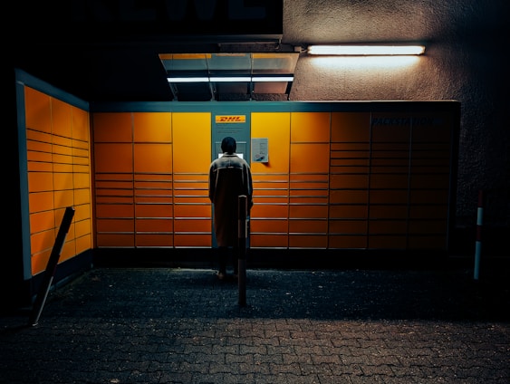 A person is standing in front of a brightly lit DHL Packstation with a grid of large yellow lockers, under a dark and slightly claustrophobic setting. The scene is illuminated by overhead lights.