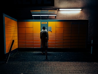 A person is standing in front of a brightly lit DHL Packstation with a grid of large yellow lockers, under a dark and slightly claustrophobic setting. The scene is illuminated by overhead lights.