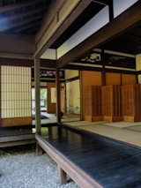 A traditional Japanese room with sliding wooden doors, tatami mats, and shoji screens. The room features wooden flooring and a tranquil setting with natural light filtering through. An open area leads to other rooms, and the wooden architecture is prominently showcased.