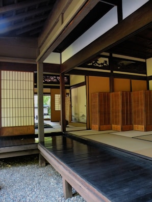 A traditional Japanese room with sliding wooden doors, tatami mats, and shoji screens. The room features wooden flooring and a tranquil setting with natural light filtering through. An open area leads to other rooms, and the wooden architecture is prominently showcased.