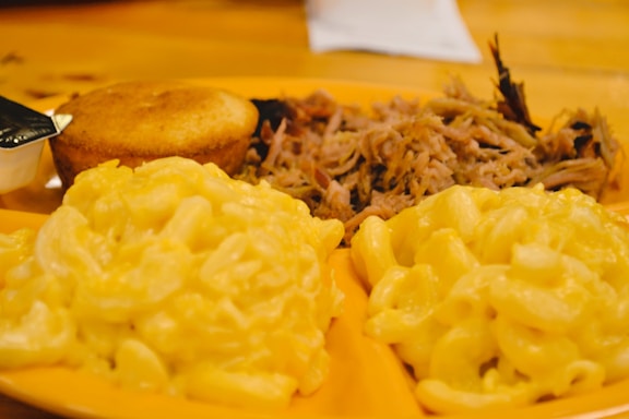 A plate with servings of creamy macaroni and cheese, cornbread, and pulled pork arranged on a wooden table.