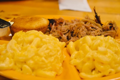A plate with servings of creamy macaroni and cheese, cornbread, and pulled pork arranged on a wooden table.