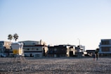 A sandy beach with a lifeguard tower numbered '10' is situated in front of several modern, multi-story beachfront houses. Two palm trees are visible near the buildings. Two people are walking on the beach. The lighting suggests either early morning or late afternoon, with long shadows and a clear sky.