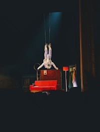 An astronaut is suspended upside down above a stage, surrounded by theatrical props including a red sofa, a floor lamp, and a backdrop with clothing hanging. The atmosphere on stage combines elements of space with a touch of theatrical whimsy.