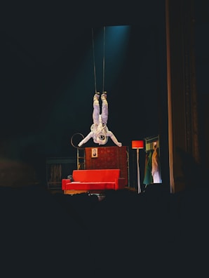 An astronaut is suspended upside down above a stage, surrounded by theatrical props including a red sofa, a floor lamp, and a backdrop with clothing hanging. The atmosphere on stage combines elements of space with a touch of theatrical whimsy.