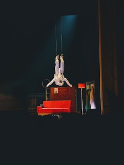 An astronaut is suspended upside down above a stage, surrounded by theatrical props including a red sofa, a floor lamp, and a backdrop with clothing hanging. The atmosphere on stage combines elements of space with a touch of theatrical whimsy.