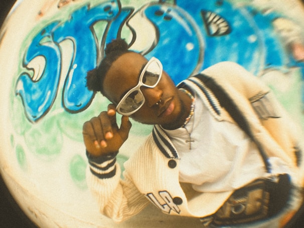 A person wearing stylish white eyewear and a cream-colored varsity jacket with black stripes is posing in front of a wall adorned with blue graffiti art. The person has their hair styled in two buns and is touching their glasses. The photo has a fisheye lens effect, giving it a distorted, artistic feel.