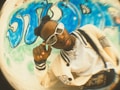 A person wearing stylish white eyewear and a cream-colored varsity jacket with black stripes is posing in front of a wall adorned with blue graffiti art. The person has their hair styled in two buns and is touching their glasses. The photo has a fisheye lens effect, giving it a distorted, artistic feel.