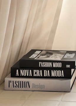 Three stacked fashion books with large, bold titles are placed near a soft, flowing curtain. The books feature grayscale covers with a modern, minimalist design.