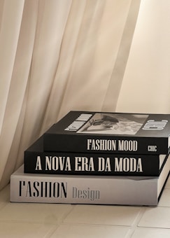 Three stacked fashion books with large, bold titles are placed near a soft, flowing curtain. The books feature grayscale covers with a modern, minimalist design.
