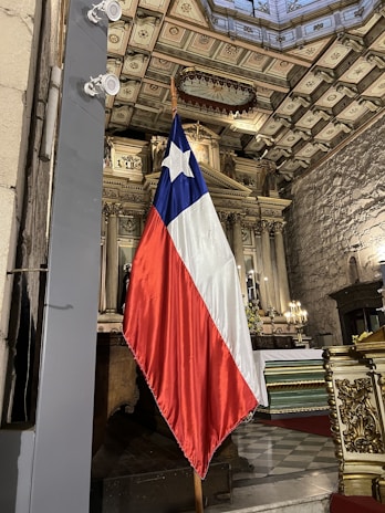 A flag with a blue field and a white star, above red and white stripes, stands inside a highly ornate church interior. The architecture includes an elaborate ceiling, intricate moldings, and decorative columns. Soft lighting and religious elements contribute to the atmosphere.