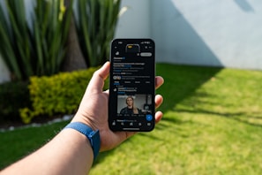 A person holds a smartphone displaying a social media profile, with a grassy lawn and tall plants visible in the background. The phone screen shows a profile with a small image of a person, and options to follow, send a message, or view posts.