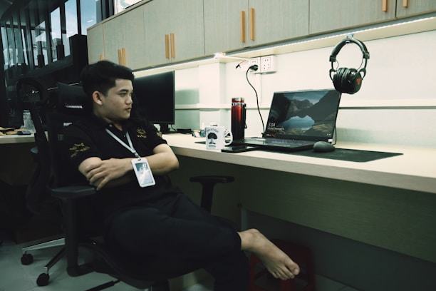 A person is sitting comfortably in an office chair, wearing a black shirt and a lanyard. The office desk has a laptop displaying a scenic landscape wallpaper, with headphones hanging nearby. There is a red insulated tumbler and a coffee mug on the desk, and the workspace has a modern setup with wooden cabinets and bright lighting.