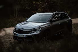 A sleek, modern SUV is parked on a dirt road surrounded by lush greenery. The vehicle's glossy finish reflects the ambient lighting, emphasizing its aerodynamic design and sophisticated front grille.