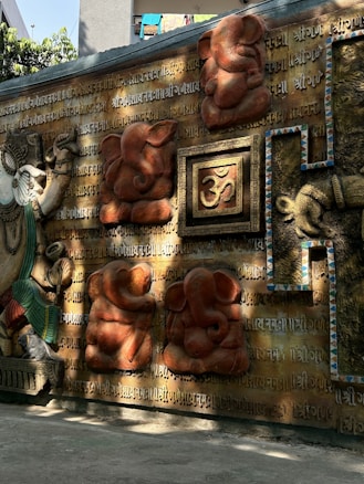 A detailed wall art featuring Hindu religious symbols and deities. The wall is adorned with intricate carvings and sculptures of Ganesha and is covered in sacred Sanskrit inscriptions. The central focus is an ornate representation of the Om symbol, framed by decorative elements.