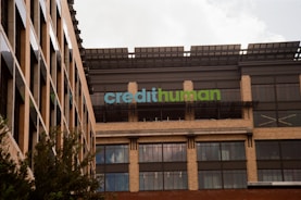 A modern building facade with large windows and a sign displaying 'credithuman'. The structure has a combination of beige brick, glass, and metal elements. Trees are partially visible in the foreground.