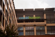 A modern building facade with large windows and a sign displaying 'credithuman'. The structure has a combination of beige brick, glass, and metal elements. Trees are partially visible in the foreground.