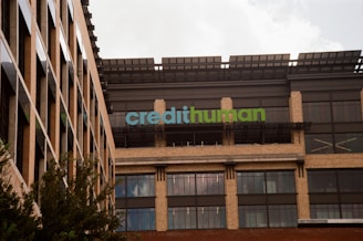 A modern building facade with large windows and a sign displaying 'credithuman'. The structure has a combination of beige brick, glass, and metal elements. Trees are partially visible in the foreground.