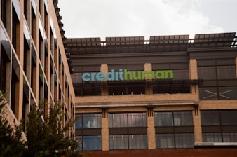 A modern building facade with large windows and a sign displaying 'credithuman'. The structure has a combination of beige brick, glass, and metal elements. Trees are partially visible in the foreground.